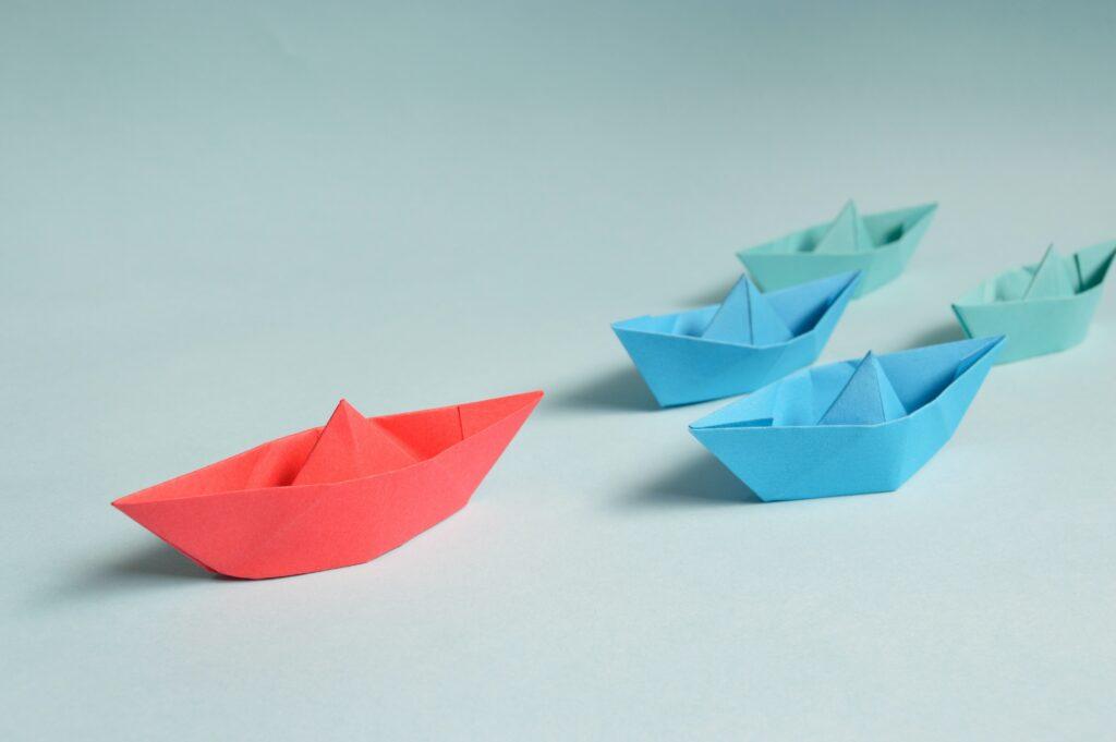 Small blue and green paper boats being lead by a red paper boat.